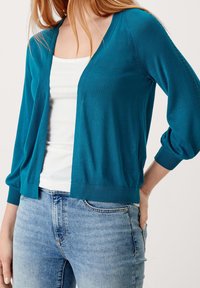 Sinisenvihreä kevyt neuletakki, jossa V-aukko, lasketut hartiat ja resorit hihansuissa, yhdistetty valkoiseen toppiin ja vaaleansiinisiin farkkuihin.