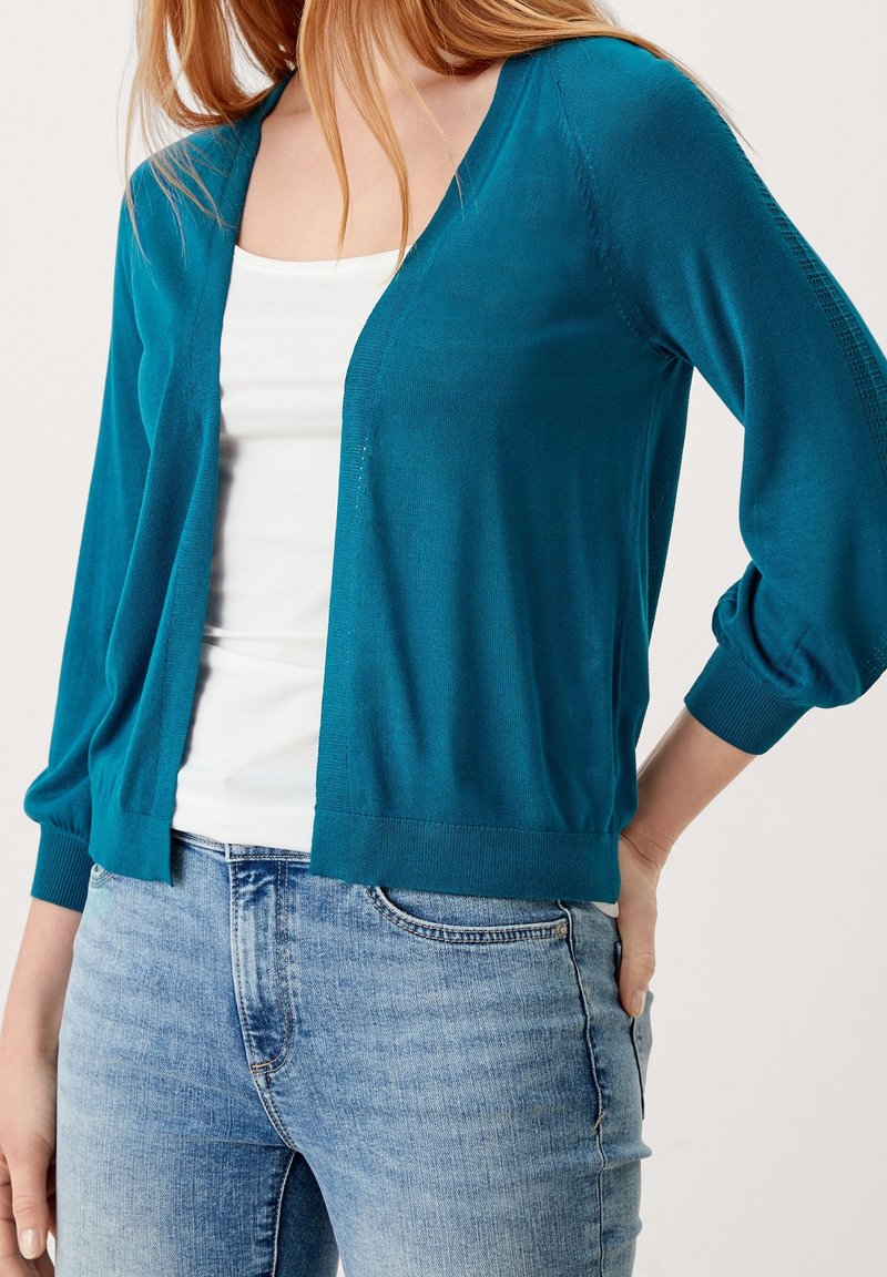 Sinisenvihreä kevyt neuletakki, jossa V-aukko, lasketut hartiat ja resorit hihansuissa, yhdistetty valkoiseen toppiin ja vaaleansiinisiin farkkuihin.