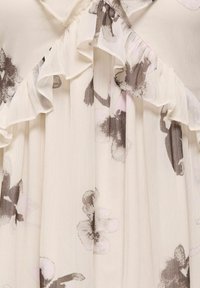 Tissu crème avec des plis doux, des imprimés floraux marron et beige, et une délicate bordure à volants le long de l'encolure.