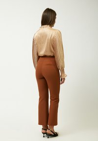 Pantaloni marroni aderenti abbinati a una blusa dorata plissettata con colletto a volant. L'outfit include sandali neri con tacco alto.