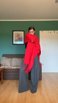 Rode gebreide sjaal met franjes, om de persoon heen gedrapeerd. Ze dragen losse grijze broeken en staan in een kamer met groene muren.