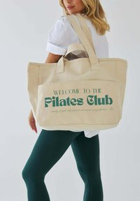 Kobieta ubrana w białą koszulkę i ciemnozielone legginsy niesie beżową torbę z napisem "Witamy w Klubie Pilates" na kieszeni.