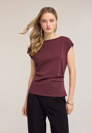 Jonge vrouw met lang blond haar draagt een bordeauxrode top met korte mouwen, zwarte broek, hangende oorbellen en een zilveren armband tegen een effen achtergrond.