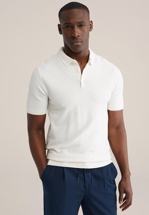 Wit gebreid poloshirt met een korte kraag, gestructureerd patroon en geribbelde mouwen, gecombineerd met marineblauwe broek.