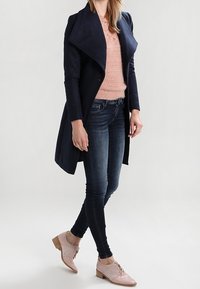 Marineblauer Wollmischmantel mit großem Kragen, hellrosa Strickpullover, dunkle enge Jeans und hellrosa Schnürschuhe mit niedrigem Absatz.