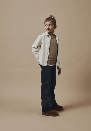 Niño vestido con una chaqueta crema texturizada, camisa de patrón marrón claro, jeans azul oscuro y zapatos marrones. El fondo es de un suave color beige.