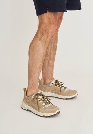 Benen met beige en witte sportsneakers met bruine veters en witte zolen, gecombineerd met marineblauwe shorts op een lichte achtergrond.