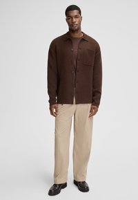 Brązowa koszula zapinana na guziki z kieszonką, noszona na brązowej bluzce, w połączeniu z jasnobeżowymi spodniami z sztruksu i czarnym obuwiem. Gładkie tło.