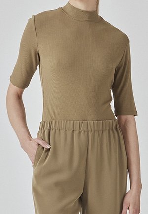 Person trägt ein eng anliegendes, kurzärmeliges, geripptes, beiges Oberteil mit hohem Kragen und lockere beige Hosen mit elastischem Bund und Taschen.