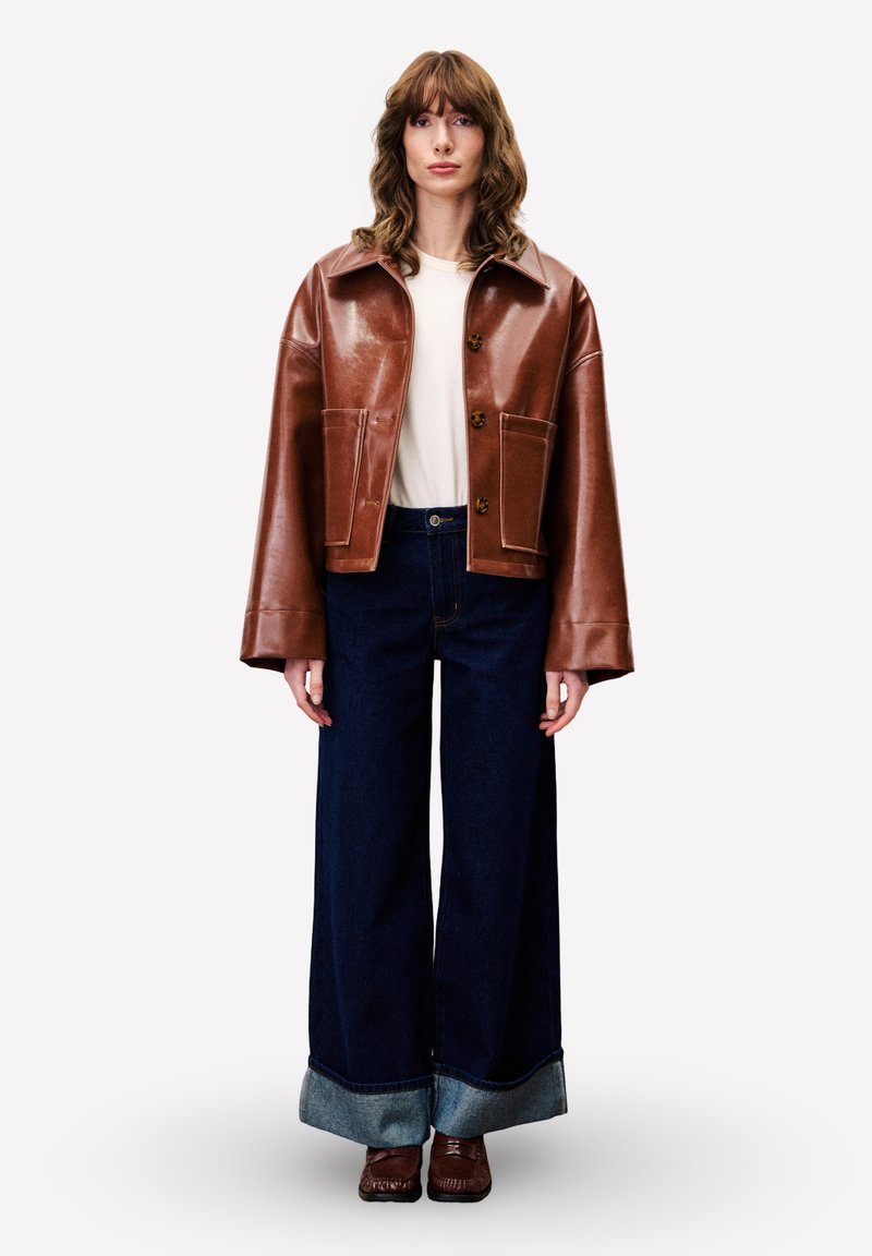 Vrouw met golvend haar, gekleed in een bruine leren jas, een witte shirt, een wijde, donkerblauwe broek met omslag, en bruine schoenen, die tegen een effen achtergrond staat.