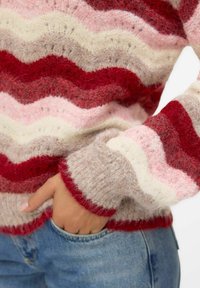 Pulover od vune s dodatkom akrila s uzorkom zigzaga u crvenoj, ružičastoj, bijeloj i bež boji, s gustim teksturom i rebrastim manžetama, nosi se preko trapera.