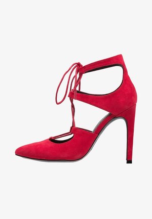 Zapato de tacón alto de ante rojo con punta afilada, laterales cortados y cordones en la parte frontal atados en un lazo alrededor del tobillo.