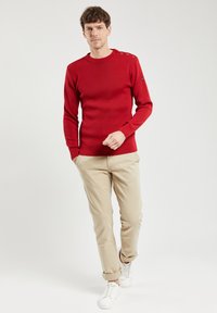 Roter Pullover mit Stehkragen aus strukturiertem Strick, mit drei Knopfakzenten an der Schulter, kombiniert mit beigen Hosen und weißen Sneakern.