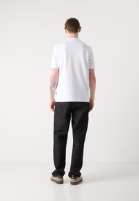 Camisa polo de manga corta blanca combinada con pantalones de pierna ancha gris oscuro y zapatillas beige, con un tatuaje sutil en el antebrazo izquierdo.