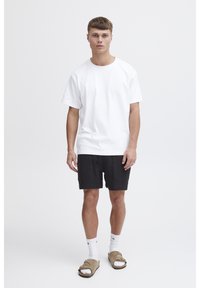 White cotton t-shirt, black shorts, and beige sandals. The shorts have a textured pattern and side pockets. Model stands against a plain background.