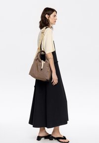 Bolso de nylon marrón con textura suave, asas cortas y una correa larga para el hombro, combinado con un vestido negro sin mangas y sandalias negras.