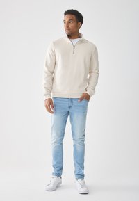 Sudadera cremallera beige claro con cuello alto, combinada con jeans azul claro y zapatillas blancas. Textura suave, corte casual.