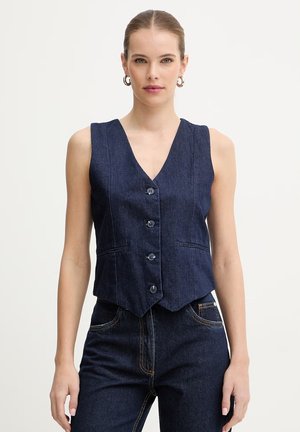 Donna che indossa un gilet di jeans blu navy senza maniche, abbinato a jeans scuri, su uno sfondo semplice.