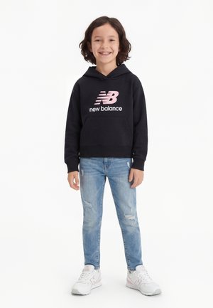 Niño sonriente con cabello rizado que lleva una sudadera con capucha negra de New Balance, vaqueros azules claros rasgados y zapatillas blancas de New Balance, de pie sobre fondo blanco.