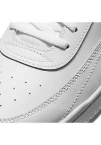 Primer plano de unas zapatillas de cuero blanco, con superficie texturizada, perforaciones y cordones planos blancos. Detalles cosidos a lo largo de los lados.