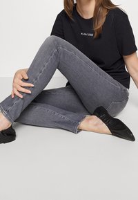 Kvinde siddende på gulvet iført en sort "MANKIND" t-shirt, grå skinny jeans og sorte spidse balletflade.