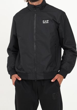 Mann trägt eine schwarze EA7 Emporio Armani Zip-Up-Jacke mit Händen in den Taschen, kombiniert mit schwarzen EA7 Emporio Armani Hosen.