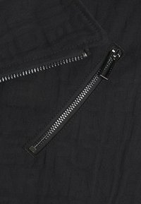 Svart tyg med subtil präglad text och två synliga silverdragkedjor, en horisontell och en diagonal.