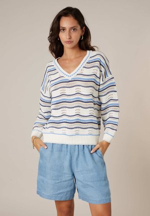 Femme portant un pull en maille blanc et bleu à rayures avec un col en V, associé à un short ample bleu clair, debout les mains dans les poches.