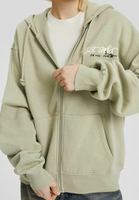 Felpa verde chiaro con zip realizzata in tessuto morbido, dotata di una tasca a canguro, polsini a coste e testo ricamato con un dettaglio grafico.