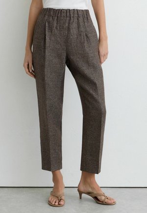 Žena nosiaca hnedé vlnkové nohavice s vysokým pásom a rovnými nohavicami s elastickým pásom a béžovými sandálmi na podpätku, stojaca opretá o bielu stenu.