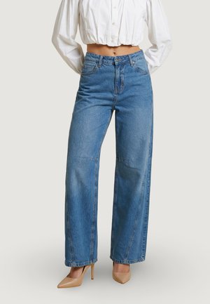 Femme portant un jean large bleu taille haute, un chemisier blanc court et des escarpins pointus beiges, debout devant un fond uni.