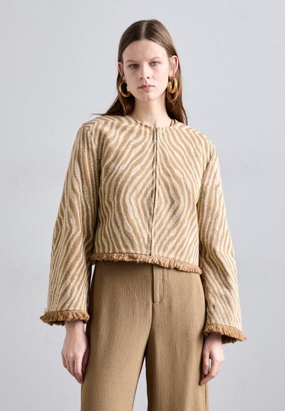 Beige kurz geschnittene Jacke mit weißem Wellenmuster, langen Ärmeln, fransigem Saum und strukturiertem Stoff. Kombiniert mit hoch geschnittenen beigen Hosen.
