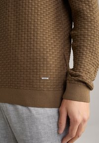 Bruine textuur trui met een geweven patroon en een geribbelde manchette. Heeft een klein wit label met "JOOP!" aan de zoom. Grijze broek zichtbaar.