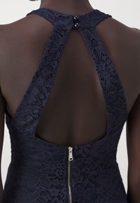 Personne à la peau foncée portant une robe en dentelle florale bleu marine avec un col licou et un dos nu, dotée d'une fermeture éclair argentée et de deux boutons.