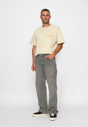 Crème katoenen t-shirt met een logo op de borst, gecombineerd met grijze cargobroeken met zijzakken en zwarte sneakers met witte zolen.