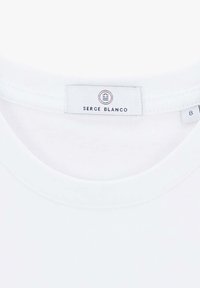 T-shirt en coton blanc avec un col rond, comportant une étiquette en tissu indiquant "SERGE BLANCO" et une étiquette de taille 8. Texture lisse, design simple.