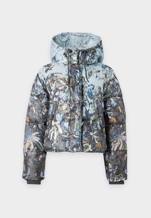 Femme portant une doudoune bleue à motifs floraux, un pull bleu clair, un jean avec une ceinture noire et des mocassins marron foncé, debout devant un fond uni.