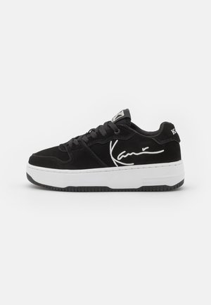 Sort lavtsiddende sneaker med hvid sål, sorte snørebånd og hvid signaturdesign på siden, vist mod en ensfarvet hvid baggrund.