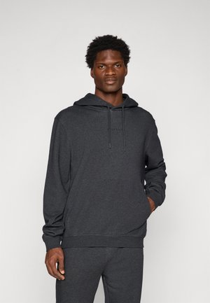 Mann in dunkelgrauem Kapuzenpulli und passender Jogginghose, steht mit einer Hand in der Tasche vor einem hellen, schlichten Hintergrund.