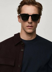Zwarte zonnebril, donker geribbeld marineblauw sweater en een bordeaux rode button-up jas met een borstzak. Schone lijnen en contrasterende texturen.