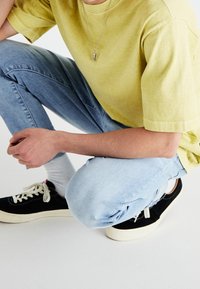 Gul bomull t-shirt, ljusblå denimjeans, svarta sneakers med vita skosnören, silvrigt hängsmycke, person som knäböjer med händerna på knäna.