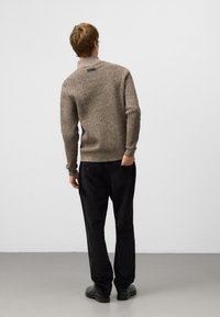 Brązowy, strukturalny sweter z wysokim kołnierzem i naszywką z logo z tyłu, w zestawie z czarnymi spodniami prostymi i ciemnymi butami.