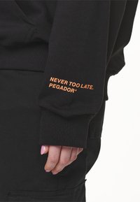Mano con uñas rosas parcialmente cubierta por la manga de un suéter negro con el texto naranja "NUNCA ES TARDE. PEGADOR*" sobre pantalones negros.