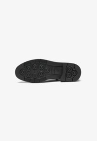 Semelle de chaussure noire avec des motifs de traction circulaires, des crampons arrondis et une surface texturée. Présente des rainures pour la flexibilité et l'adhérence.
