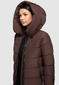 Veste en duvet marron avec grande capuche, fermeture zippée et poches latérales. Présente une texture matelassée et des accents de fermeture éclair noirs.