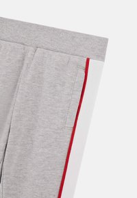 Graue Jogginghose mit geripptem Bund, seitlichen Taschen und roten sowie weißen Akzenten an den äußeren Nähten. Aus weichem Baumwollmaterial.