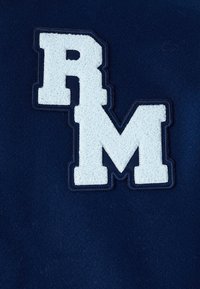 Biała, teksturowana naszywka z literami „RM” przyszyta na ciemnoniebieskim tle z tkaniny.