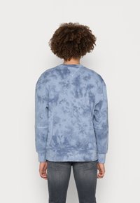 Persona con cabello rizado, vistiendo un sudadera azul con tie-dye y jeans gris oscuro, de pie frente a un fondo claro y liso.