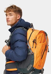 Pomarańczowy i szary plecak z regulowanymi paskami, zamkami błyskawicznymi i wieloma przegródkami, noszony przez osobę w granatowej kurtce puchowej.