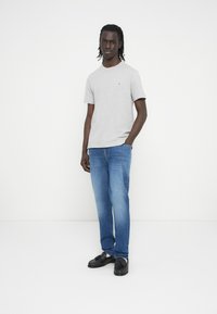 Γκρι κοντομάνικη μπλούζα με μικρό λογότυπο, σε συνδυασμό με ανοιχτό μπλε τζιν και μαύρα παπούτσια. Το μοντέλο στέκεται μπροστά σε απλό φόντο.